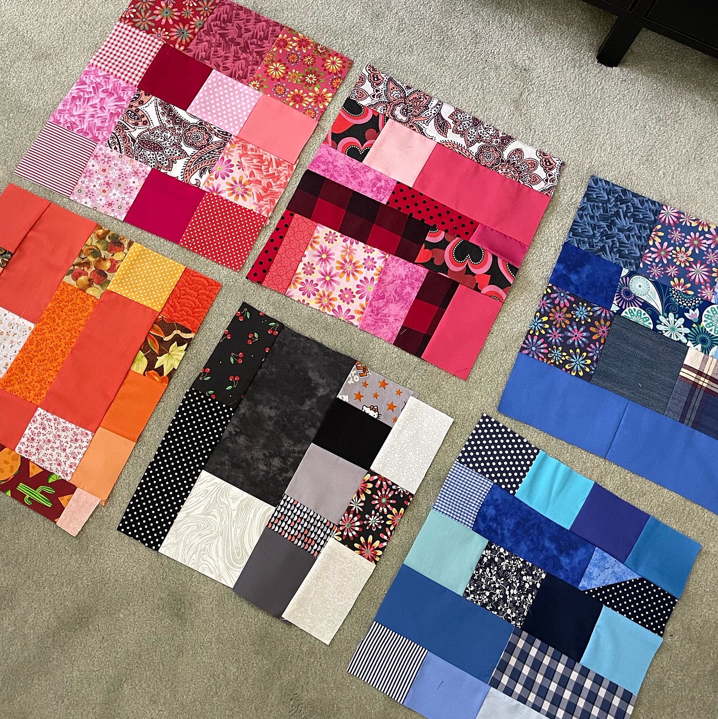 An assortment of colorful quilt blocks laid out on a light carpeted floor. Each block is composed of various fabric squares and rectangles in distinct color themes: one in shades of red and pink, another in orange and yellow hues, one with a mix of black, white, and gray fabrics, and two in different tones of blue. Patterns include floral prints, stripes, plaids, and solids, adding texture and visual interest to the patchwork designs.
