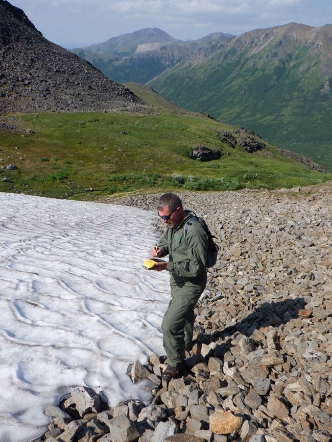 An archeologist stands on the edge of a snow patch, where it meets rocky ground, and uses a hand-held GPS device.