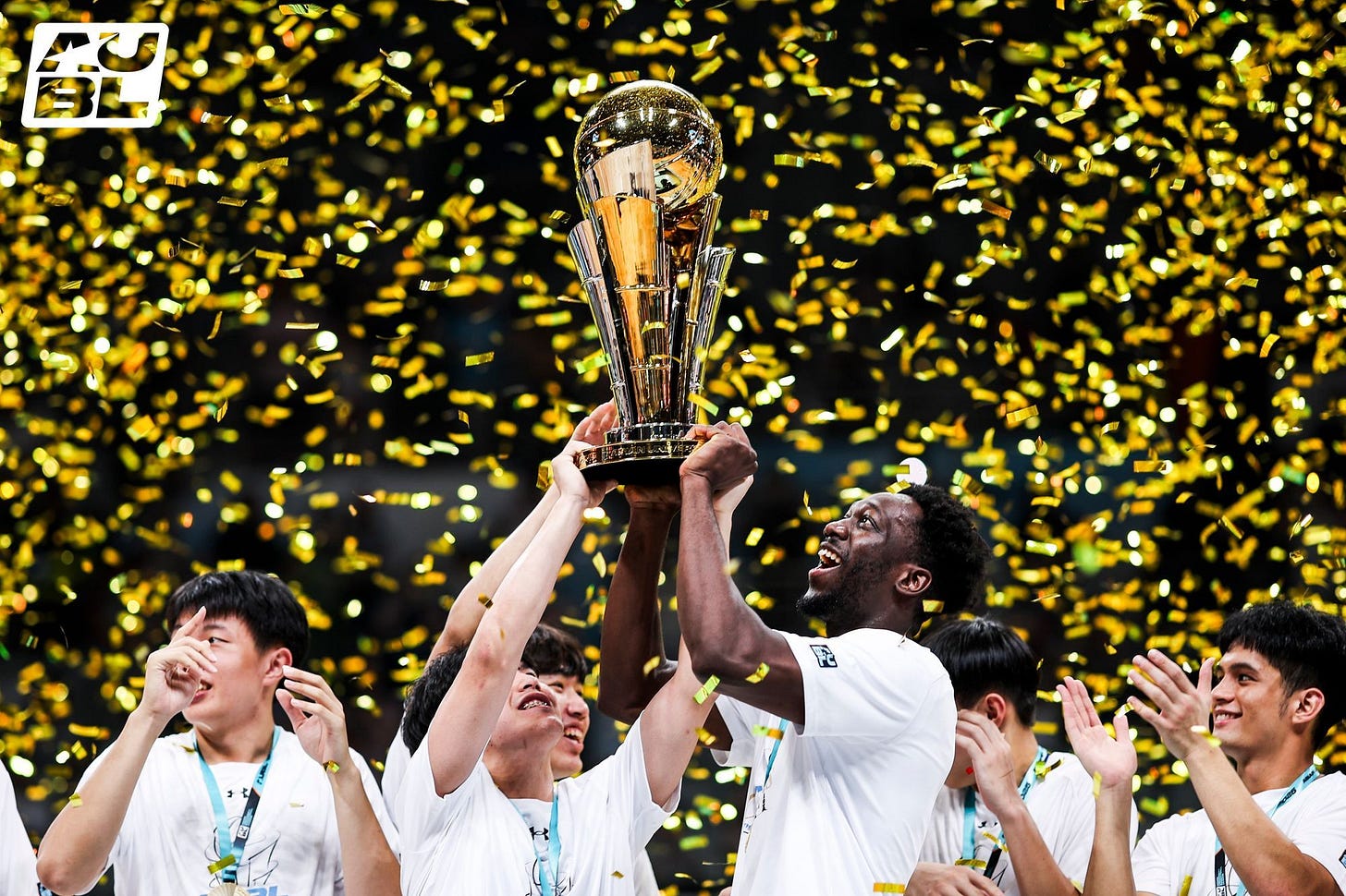 basketball players from National Chengchi University (NCCU) celebrate as they lift a tall, gleaming gold championship trophy into the air amidst a heavy shower of glittering gold confetti. The players wear white championship t-shirts, medals around their necks, and look up with expressions of pure joy and excitement. An "AUBL" logo is visible in the top left corner, representing the Asian University Basketball League.