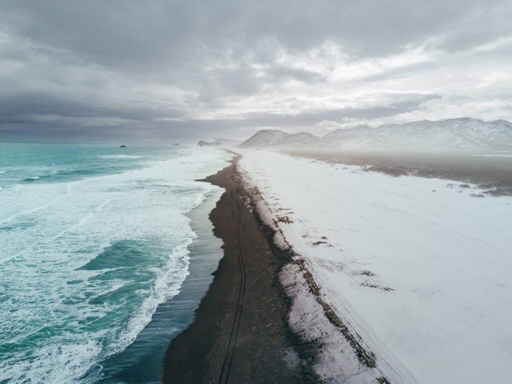 The Wisdom of WIntering - aerial view of snowy beach meeting cold ocean waves, moody gray sky and mountains in distance