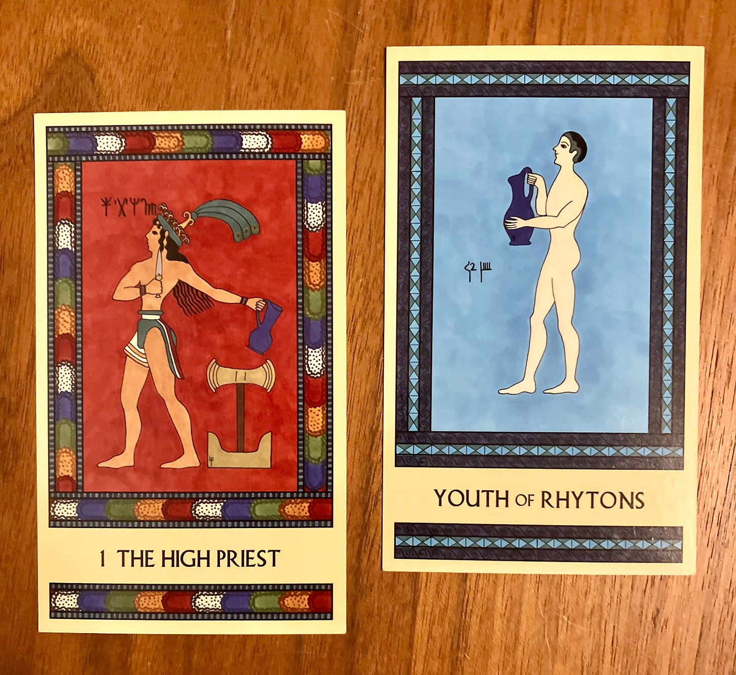 Two Minoan Tarot cards side by side on a brown wood surface. The High Priest has a bright multicolor border and a dark red background. It shows a Minoan man wearing a loincloth and a feathered hat, striding toward the left. He is holding a dagger and a pitcher, while a labrys and a pair of sacred horns stand by his feet. The Youth of Rhytons is painted in shades of blue. It shows a Minoan boy, naked, standing and facing left. He is holding a large blue pitcher. 