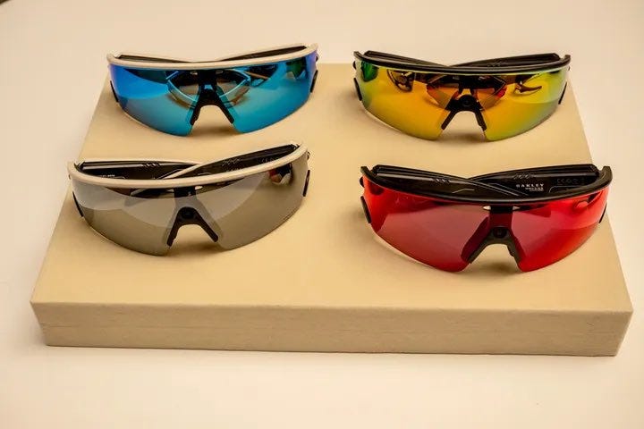Four pairs of sunglasses arranged on a beige rectangular box. The sunglasses include Ray-Ban and Oakley Vanguards models, each with distinct colored lenses: blue, silver, orange, and red. A centered camera is visible on the Oakley Vanguards.