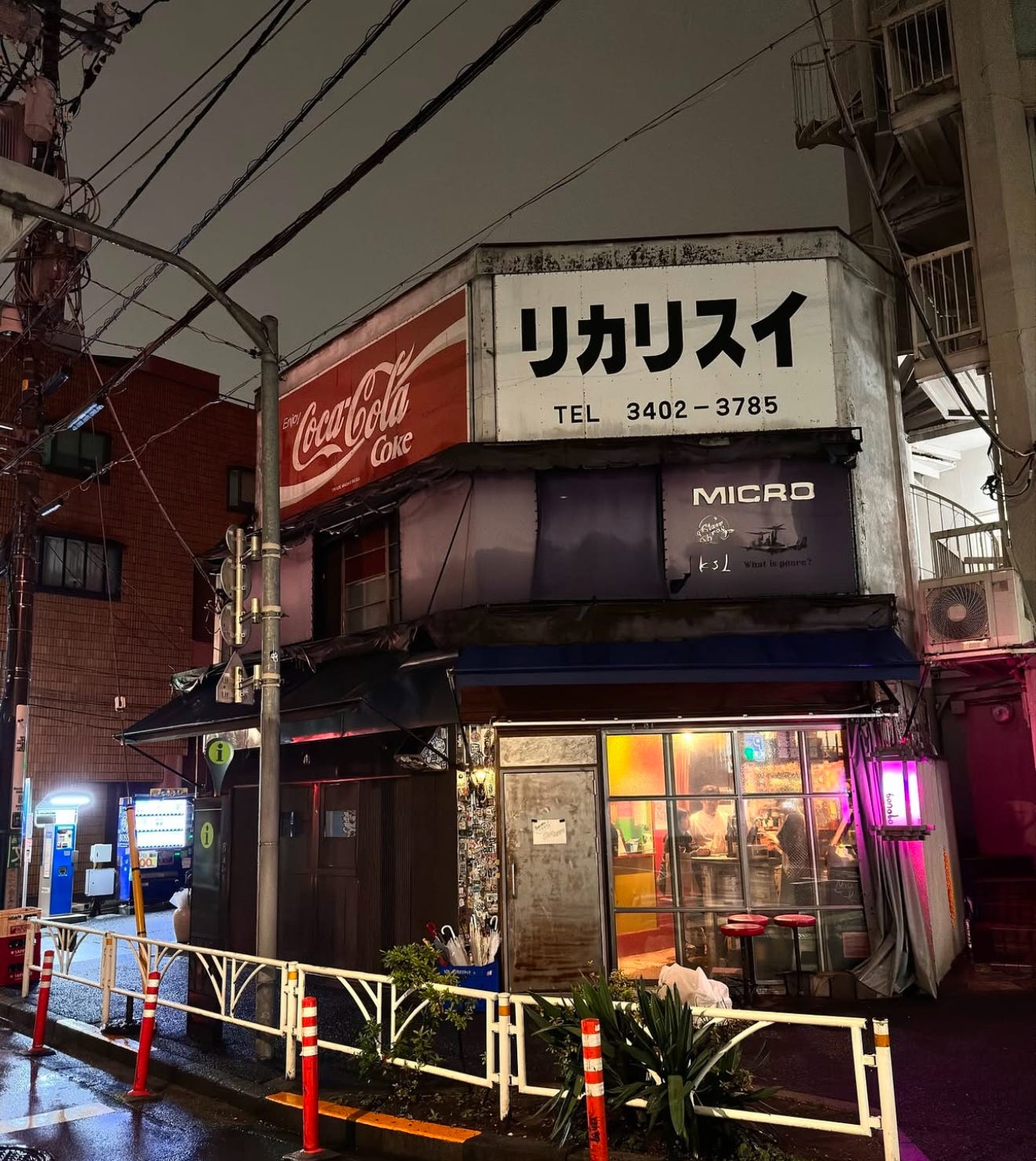A cozy, dimly-lit bar on a rainy Tokyo street corner, glowing with warm interior lights and a neon sign. The exterior features vintage Coca-Cola signage and Japanese text, giving it a nostalgic, retro vibe. A couple of stools and umbrellas rest outside, while patrons can be seen inside enjoying drinks and conversation.