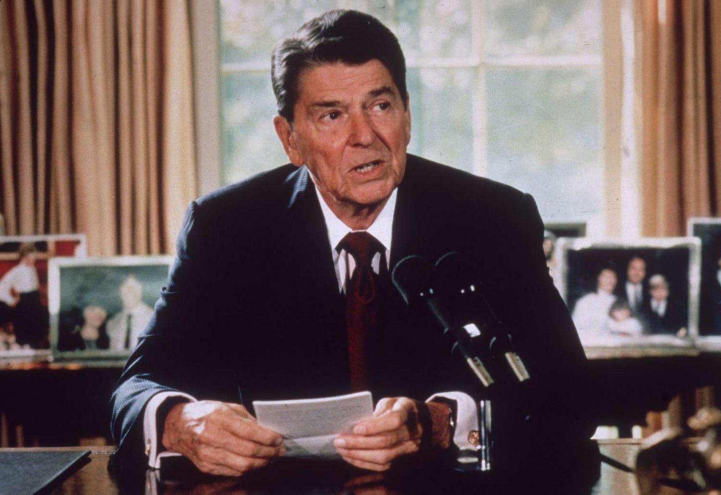 Ronald Reagan speaks into microphones at his desk, holding notes, with family photos behind him. Ronald Reagan speaks into microphones at his desk, holding notes, with family photos behind him.