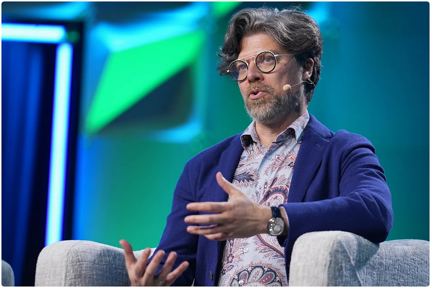 Bryan Catanzaro, VP of Applied Deep Learning at Nvidia, gesturing mid-sentence during an on-stage interview with Axios at GTC 2026. He wears glasses, a navy blazer over a patterned shirt, and a headset microphone. The stage is lit in blue and green.