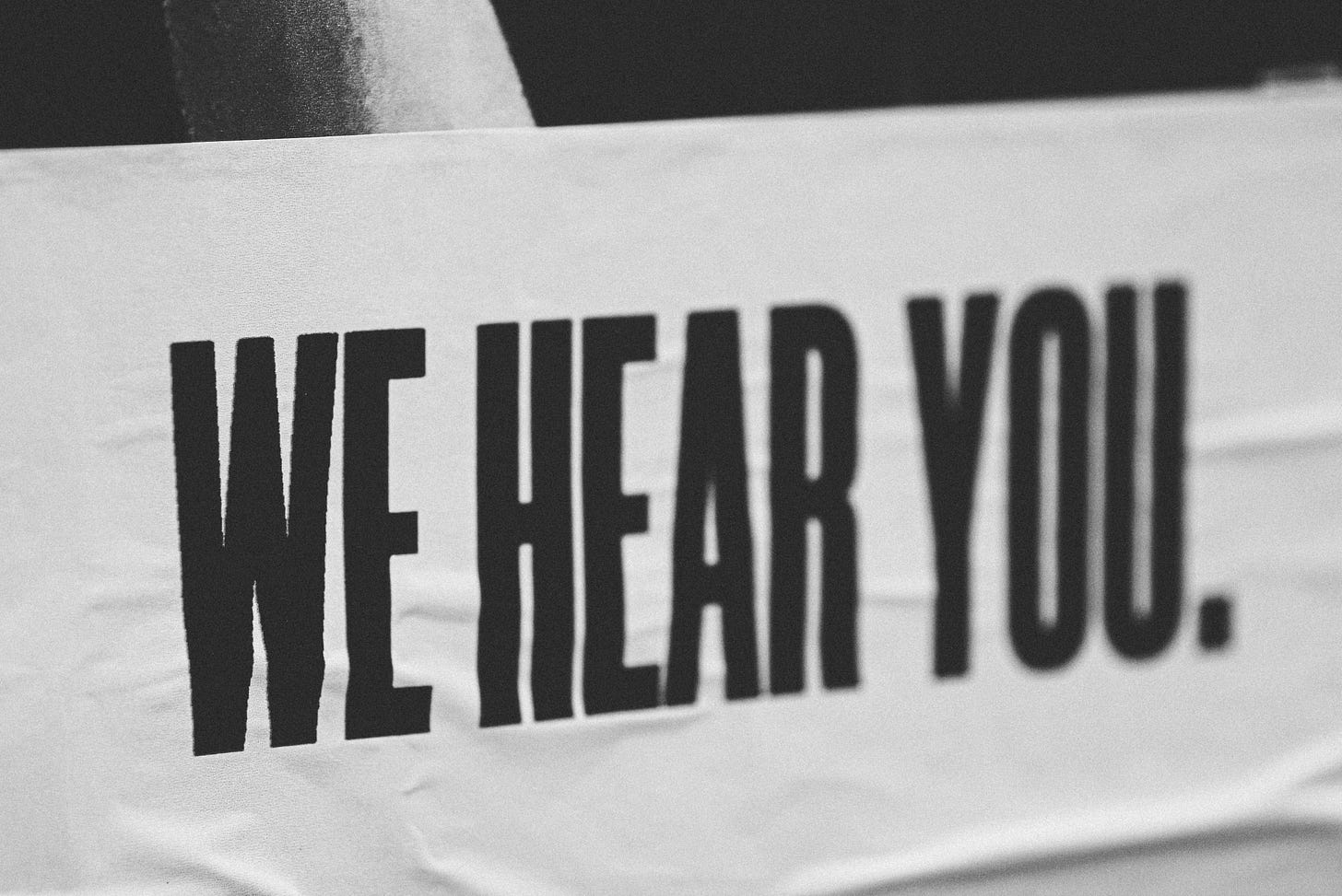 A black and white photo of a sign with a white background and the words "We Hear You" all capitalized and in black.