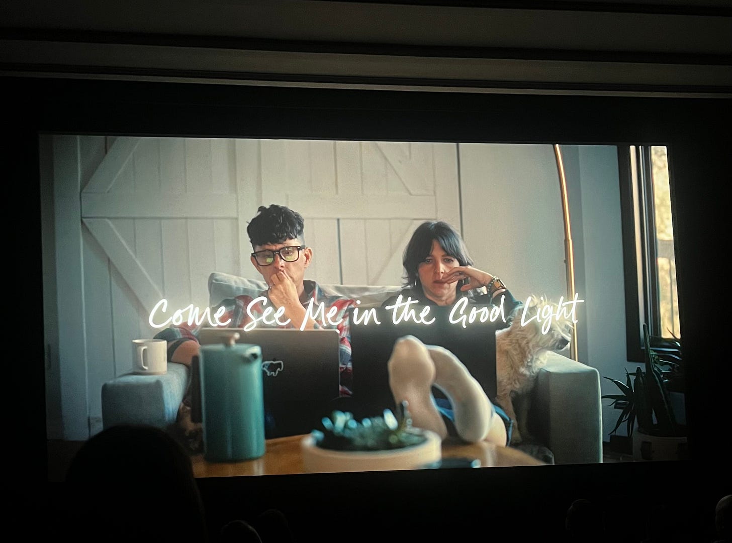 movie screen projecting Andrea and Meg, sitting in a single love-seat with computers on both of their laps, writing together. French-press coffee pot and mugs in the shot. "Come See Me in the Good Light" in a neon cursive across their bodies. movie screen projecting Andrea and Meg, sitting in a single love-seat with computers on both of their laps, writing together. French-press coffee pot and mugs in the shot. "Come See Me in the Good Light" in a neon cursive across their bodies.