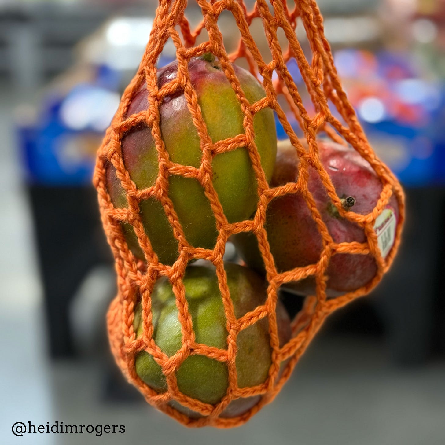 Orange, hand-crocheted net bag holds three mangoes in front of a blurry background of the produce section at the supermarket.