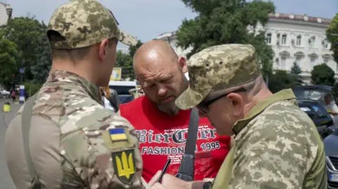Thanyarat Doksone/BBC Conscription officers Anatoliy (in sunglasses) and Oleksiy stop a man on the streets of Odesa Thanyarat Doksone/BBC Conscription officers Anatoliy (in sunglasses) and Oleksiy stop a man on the streets of Odesa