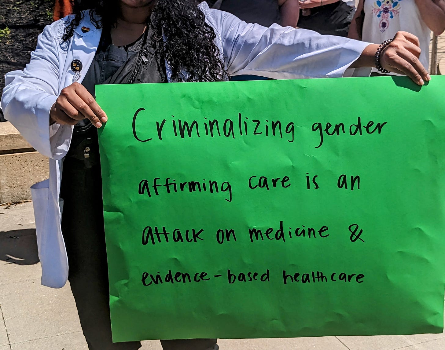 A photograph of a medical student in a doctor's coat holding a green sign that reads "Criminalizing gender-affirming care is an attack on medicine & evidence-based healthcare."