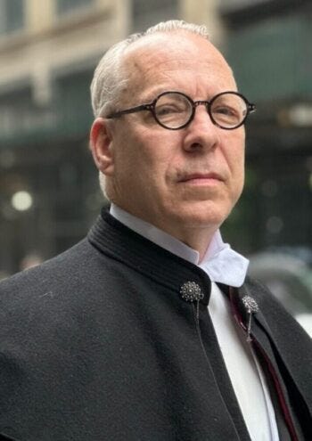 Jeffrey Tucker in round glasses and a dark high collared coat looks into the distance on a city street. Jeffrey Tucker in round glasses and a dark high collared coat looks into the distance on a city street.