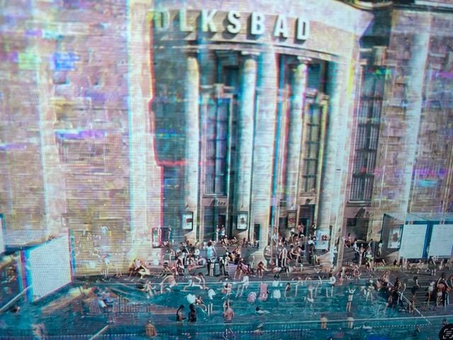 Eine große historische Gebäudefassade mit der Aufschrift „VOLKSBAD“ und hohen Säulen. Vor dem Eingang befindet sich ein temporäres Wasserbecken, in dem zahlreiche Menschen baden, sitzen und sich aufhalten. Eine große historische Gebäudefassade mit der Aufschrift „VOLKSBAD“ und hohen Säulen. Vor dem Eingang befindet sich ein temporäres Wasserbecken, in dem zahlreiche Menschen baden, sitzen und sich aufhalten.