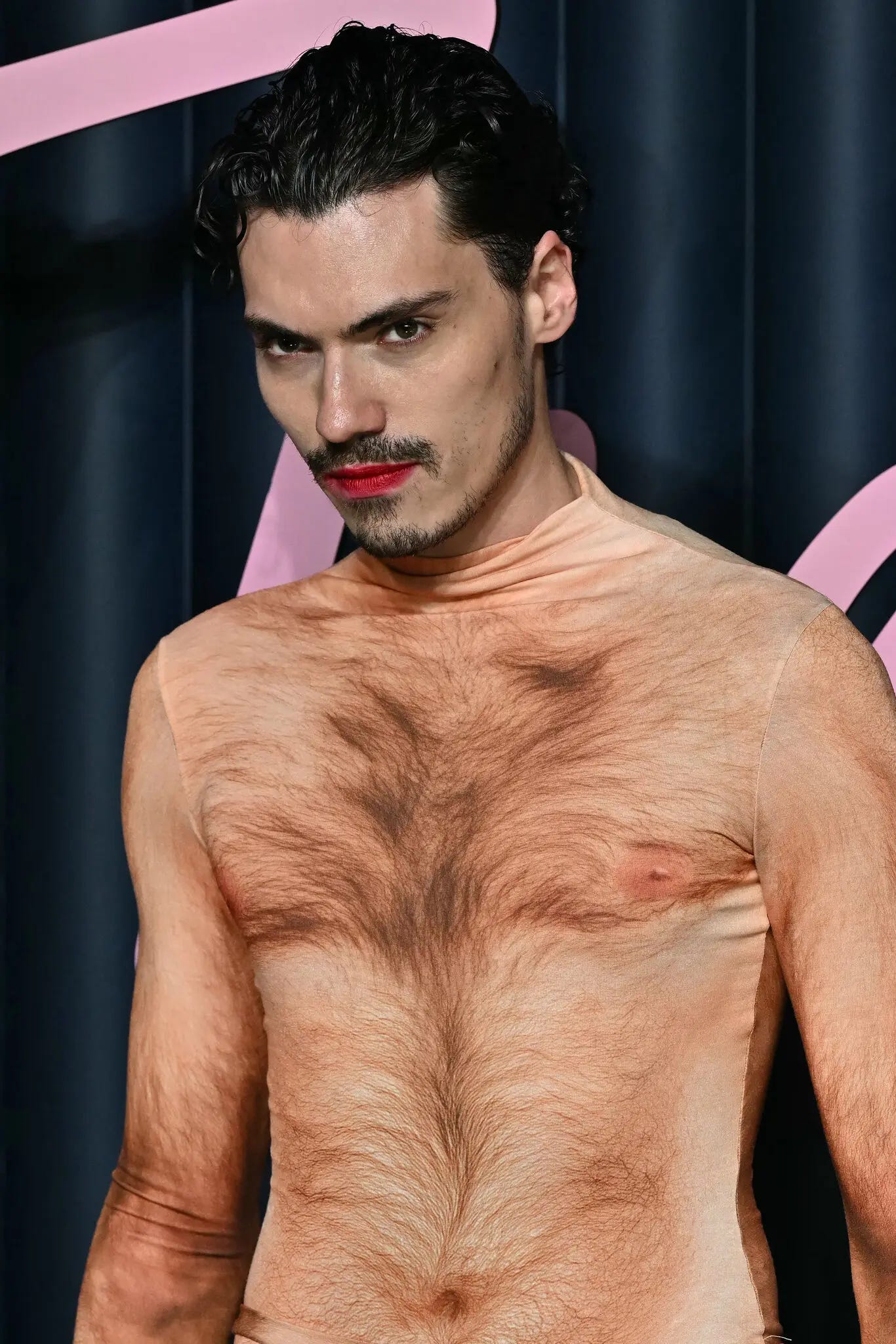 Fashion influencer Elias Medini wears a form-fitting bodysuit printed with a hyper-realistic image of a nude, hairy male torso. He stares intensely at the camera, lips painted red, against a dark backdrop with pink lettering.