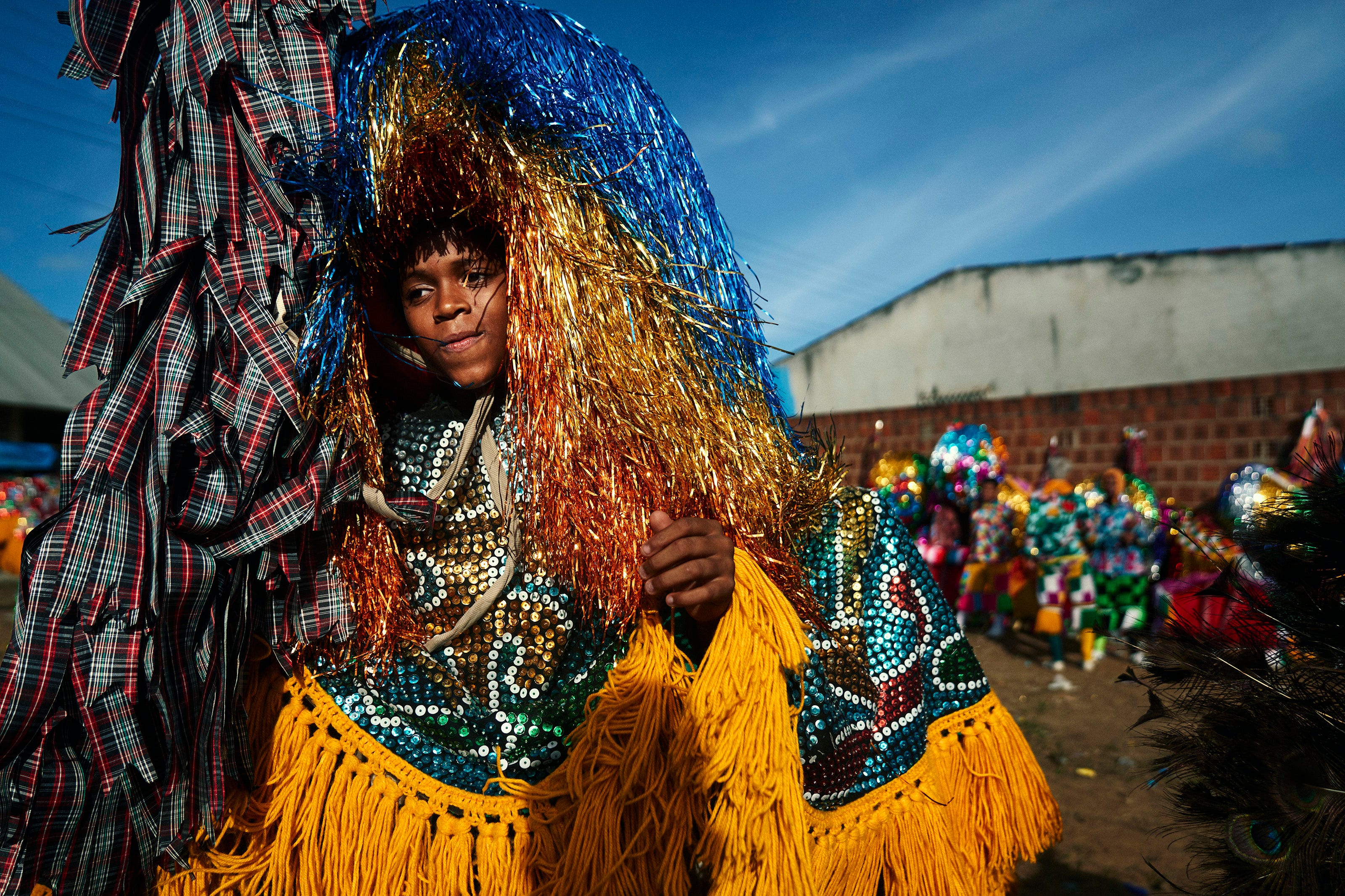 A person in a vibrant, colorful costume with sequins and long metallic tinsel stands outdoors at a festival, surrounded by bright decorations and a blue sky in the background.