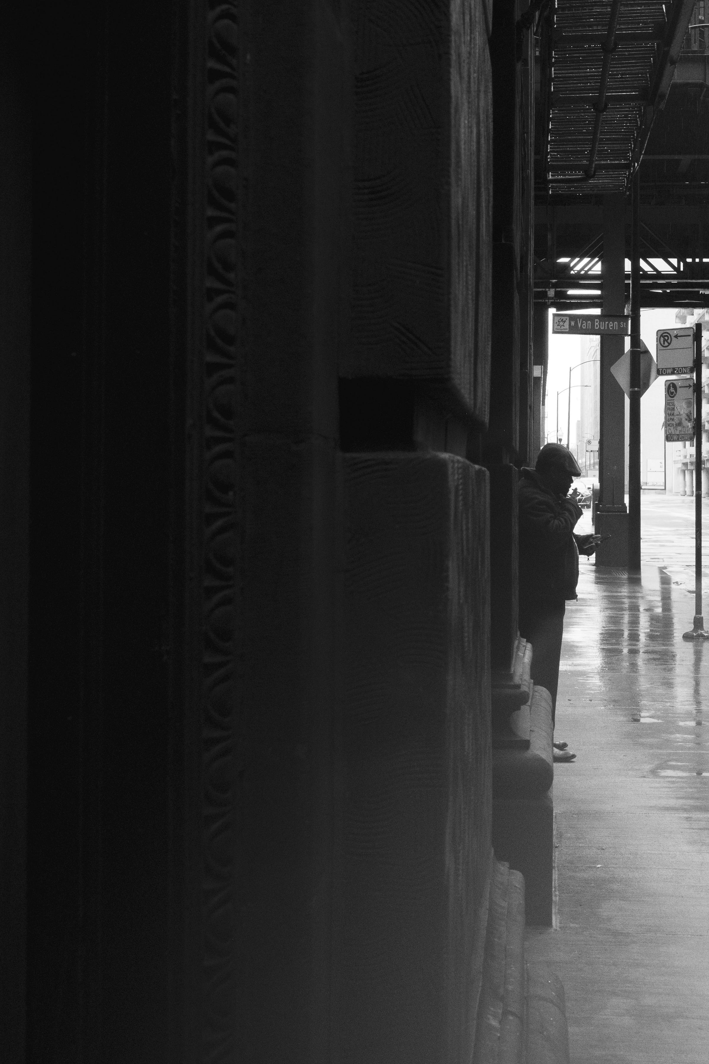 A person stands under an overpass on a rainy day, smoking and looking at their phone, framed by the textured facade of a historic building. Reflections on the wet sidewalk add to the moody atmosphere. A person stands under an overpass on a rainy day, smoking and looking at their phone, framed by the textured facade of a historic building. Reflections on the wet sidewalk add to the moody atmosphere.