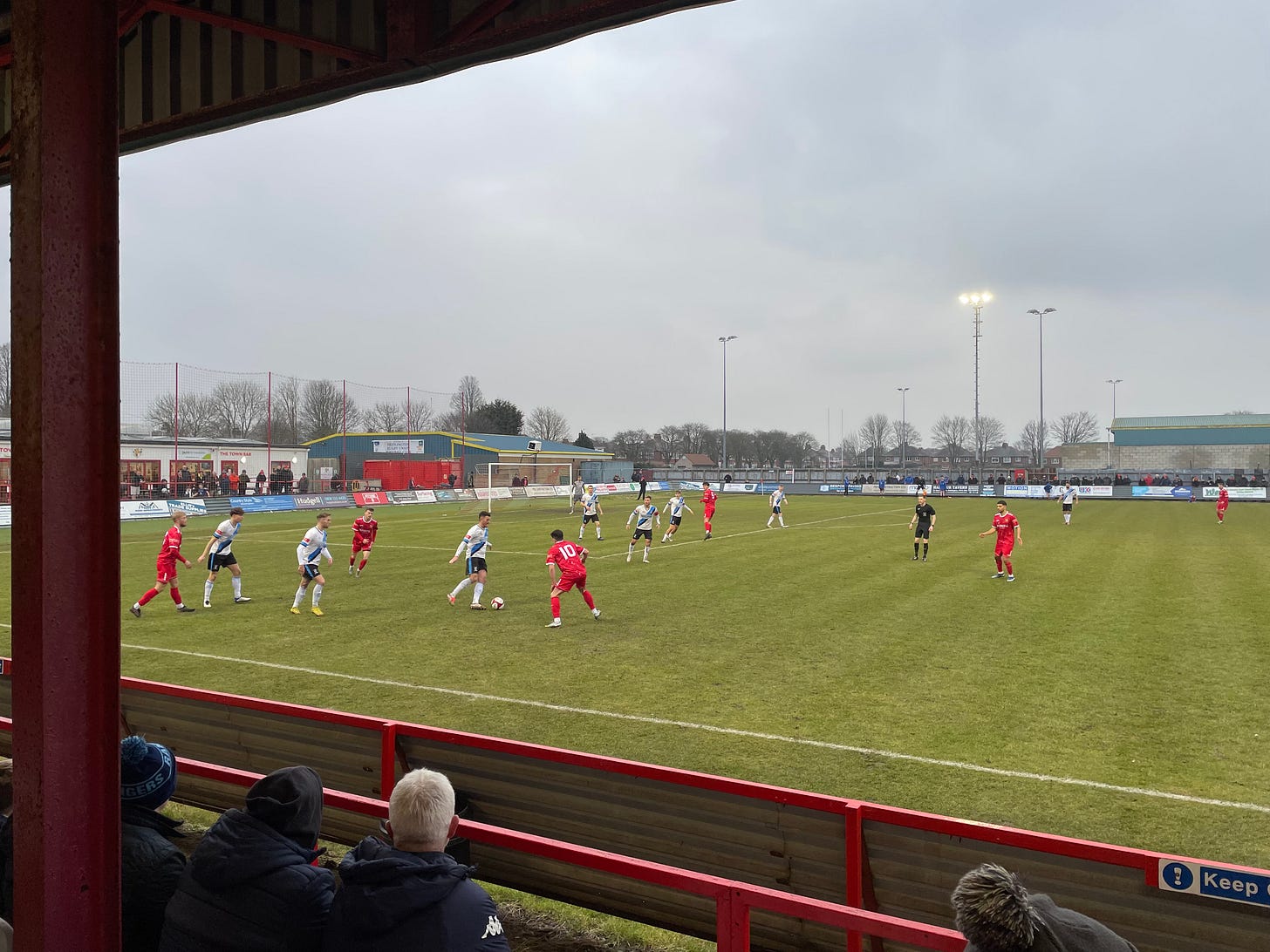 The game is in progress. Looking out from the stand, framed by the stanchions and roof, we look towards the goal with players in white and red on the pitch.