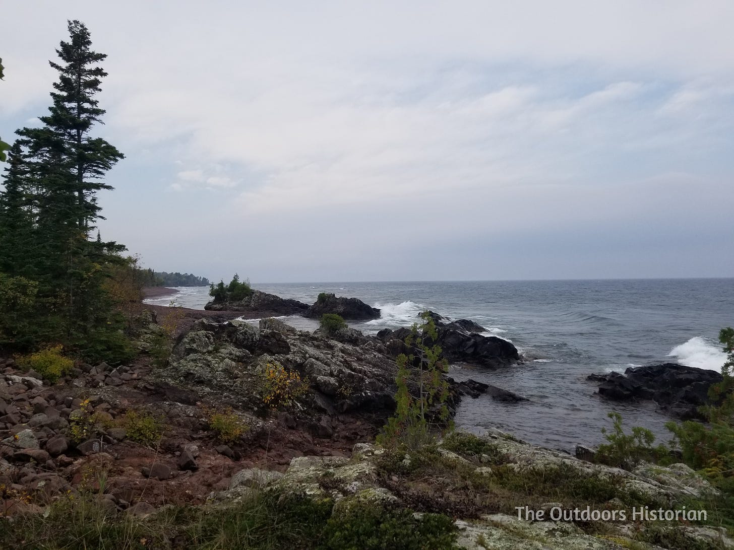 A rocky section of coast with the occasional pine tree on a grey day.The rocks illustrate the tilted layers of the eastern end of the trough formed by the Mid-Continental Rift.