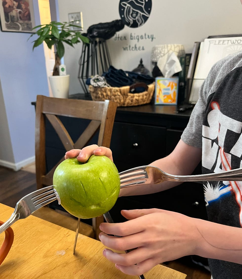 A picture of a child's hand holding up a green apple with a smiley face and eyes carved into it, a fork on each side to make arms, and two butter knives stuck on the bottom to make legs.