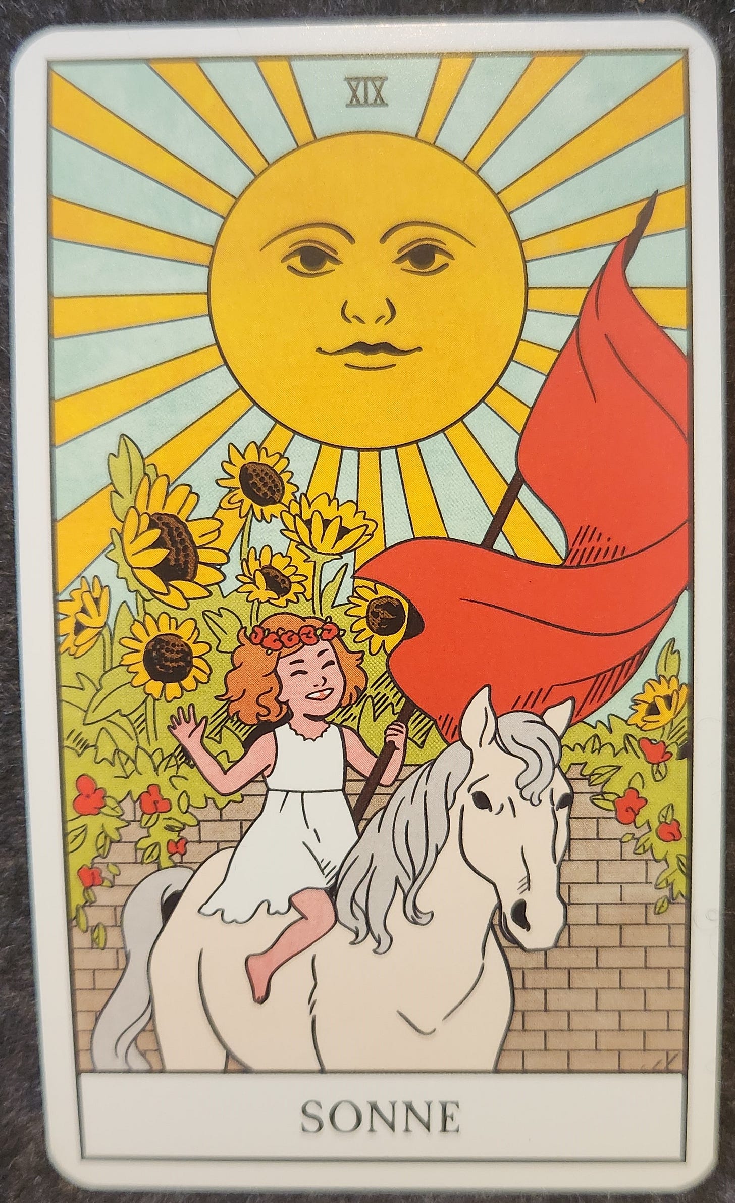 A tarot card labelled XIX Sonne. A small smiling child is riding on the back of a white horse. She is dressed in a white dress and carrying a red flag. She is smiling up to a brightly shining sun. Behind her is a garden of sunflowers. 