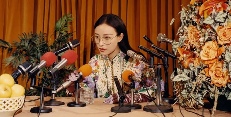This may contain: a woman sitting at a table with microphones in front of her and fruit behind her
