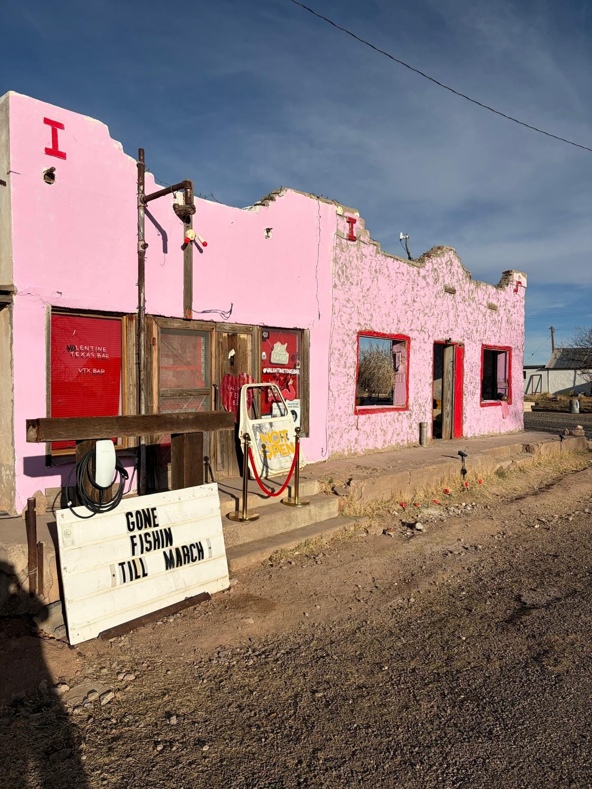 The knackered pink facade of Valentine Texas Bar stands roadside with clear blue desert skies behind. There's a sign saying "gone fishin till march" out front