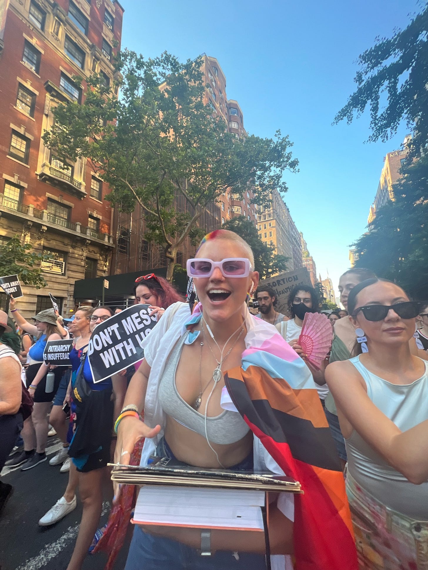 Kat when they ran into their first Dyke March. They are smiling at the camera while holding thrifted books and records. Behind them is a crowd of queer people walking down 5th avenue. 