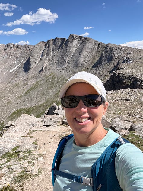 wildflowers along a rocky mountain trail; a woman standing in front of a view of mount blue sky; a mountain goat