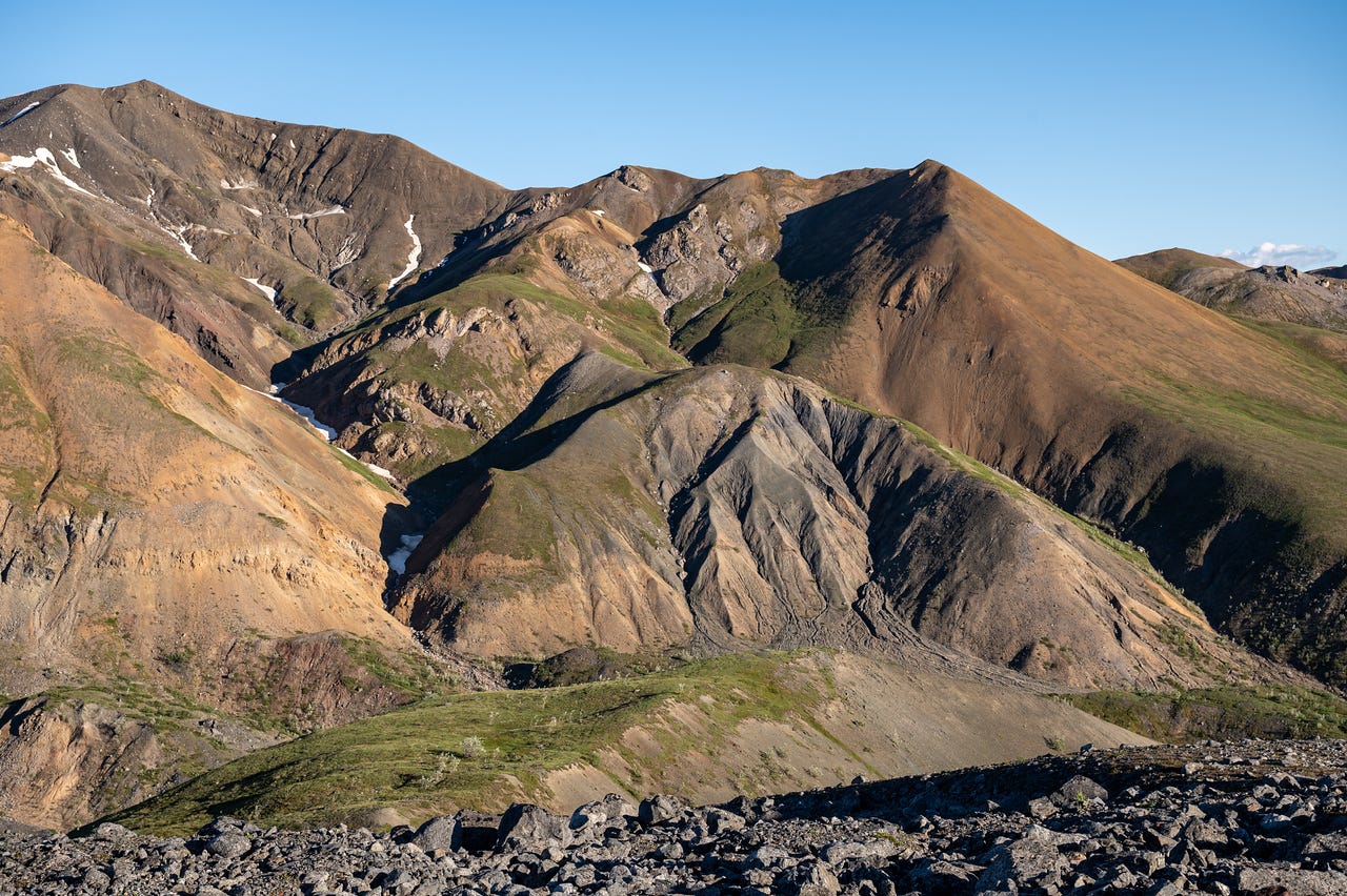 Layered ridges of rust-red, ochre, and grey rock rise across the frame, their lower slopes streaked with green tundra and cut by steep gullies, a small snow patch lingering in one shaded draw. A field of dark talus fills the foreground under a clear blue sky.