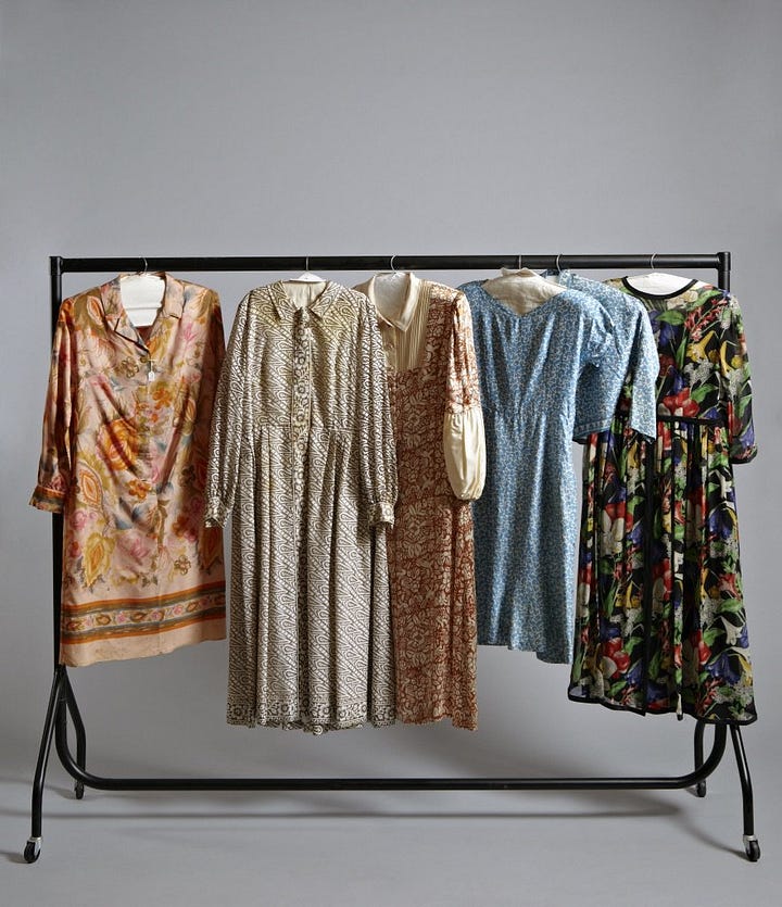 1) Classic black-and-white portrait of Gluck in a loose white shirt/smock, looking down and to the side. Their expression is thoughtful, with shadows adding depth. They have short dark hair, and they are holding a cigarette in their right hand. There is a dark shirt sticking out from under the larger shirt, with both collars sticking up. 2) A white mannequin displays a vintage, cream-colored linen shirt/smock with a black collar and buttoned cuffs, evoking a classic, timeless style. It's positioned in the same style Gluck was wearing it in the previous photo, including the collars sticking up. The mannequin head is also looking to the side, like Gluck was. 3) A pair of vintage brown leather shoes with buckles and low heels on a neutral background, conveying a sense of timeless style and craftsmanship. 4) Five vintage dresses/gowns hang on a black clothing rack with a small jacket. Patterns include floral, paisley, and abstract designs, evoking a nostalgic and elegant feel. 5) A white mannequin displays an elegant black evening gown with a lace overlay. The dress features long sleeves and a floor-length skirt, conveying sophistication.