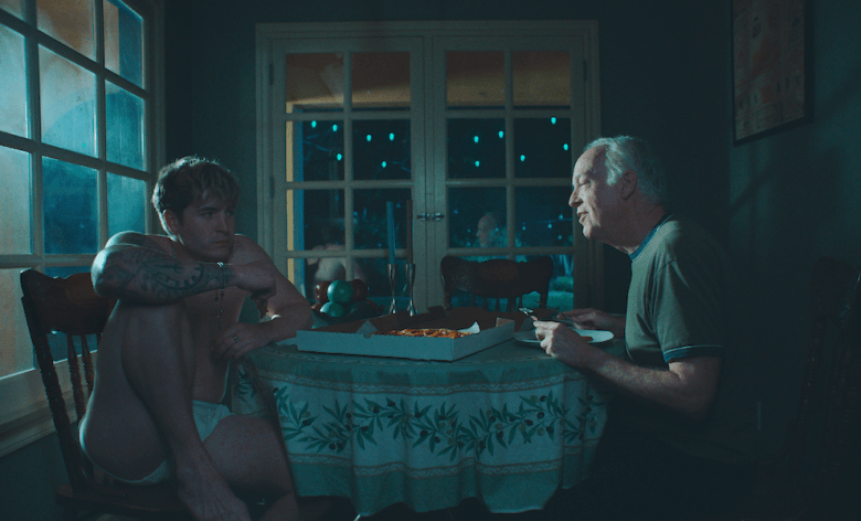 In a still from the movie Blue Film, a young man sits on the left side of the frame in a chair, with his knees pulled up. Across the table sits an older man. 