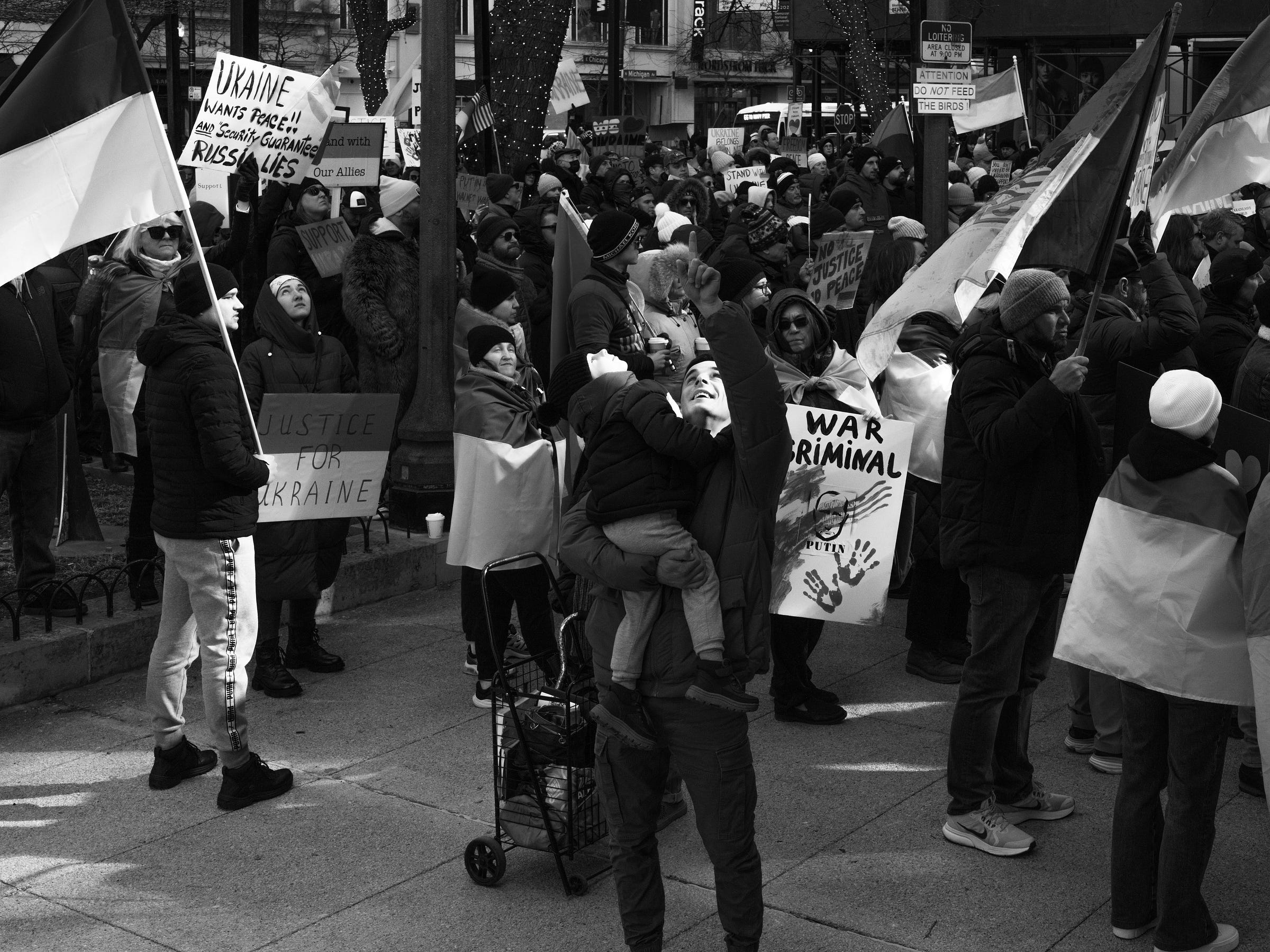 A black and white photograph captures a large protest in support of Ukraine. The crowd, bundled in winter clothing, holds various signs and flags. A prominent sign reads "Justice for Ukraine," while another declares "War Criminal" with an image of Vladimir Putin. A man in the center lifts a child onto her shoulders as they reach upward, surrounded by protesters waving Ukrainian flags. The scene is filled with determined expressions and a strong sense of collective resistance. A black and white photograph captures a large protest in support of Ukraine. The crowd, bundled in winter clothing, holds various signs and flags. A prominent sign reads "Justice for Ukraine," while another declares "War Criminal" with an image of Vladimir Putin. A man in the center lifts a child onto her shoulders as they reach upward, surrounded by protesters waving Ukrainian flags. The scene is filled with determined expressions and a strong sense of collective resistance.