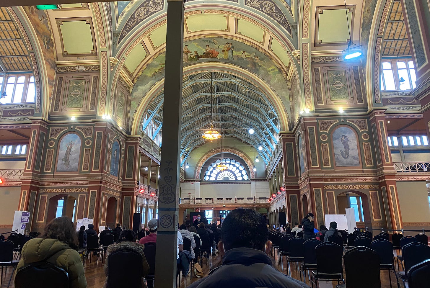 The left image shows a mural on the ceiling of Melbourne's Royal Exhibition Building called 'The Arts Applied to Peace', with people sitting in rows below waiting for their Covid jabs. The right image is a close up of the same mural, covered up, under restoration. The left image shows a mural on the ceiling of Melbourne's Royal Exhibition Building called 'The Arts Applied to Peace', with people sitting in rows below waiting for their Covid jabs. The right image is a close up of the same mural, covered up, under restoration.