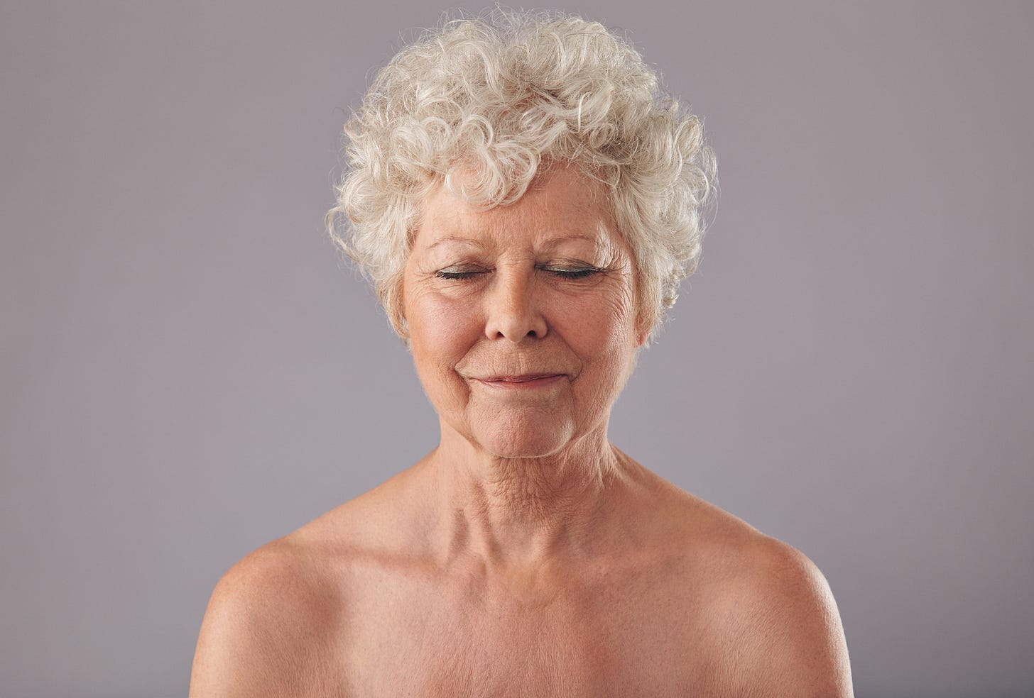 Older woman with short curly white hair standing bare-shouldered against a plain background, eyes closed and a peaceful smile on her face.