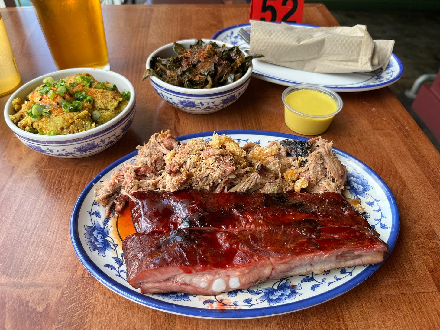 A large plate with spare ribs, glazed with wine red sauce, and pulled pork. Small bowls with yellow curry cornbread with garnish on top, and collard greens with red spices on top. 