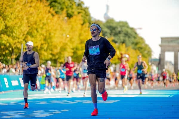 Harry Styles, in a dark long-sleeve shirt, black shorts and orange shoes, runs on a blue surface.
