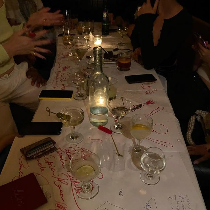 This may contain: a group of people sitting at a long table with wine glasses on top of it