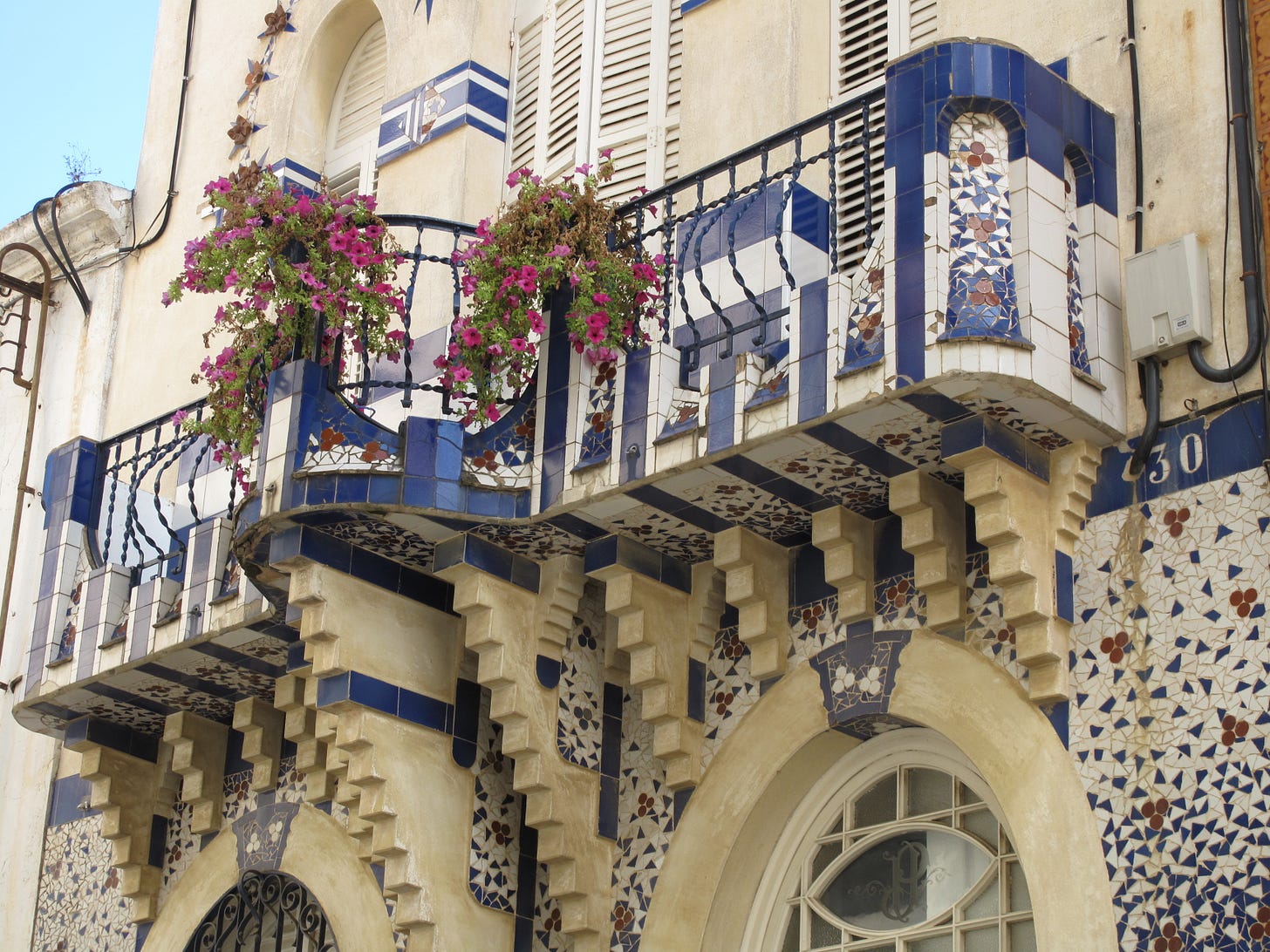 Dettaglio del balcone di Can Planiol a Sant Pol de Mar, un edificio bianco con facciate decorate di mosaici bianchi e azzurri. Sul balcone, due vasi di fiori fucsia aggiungono colore alla facciata.