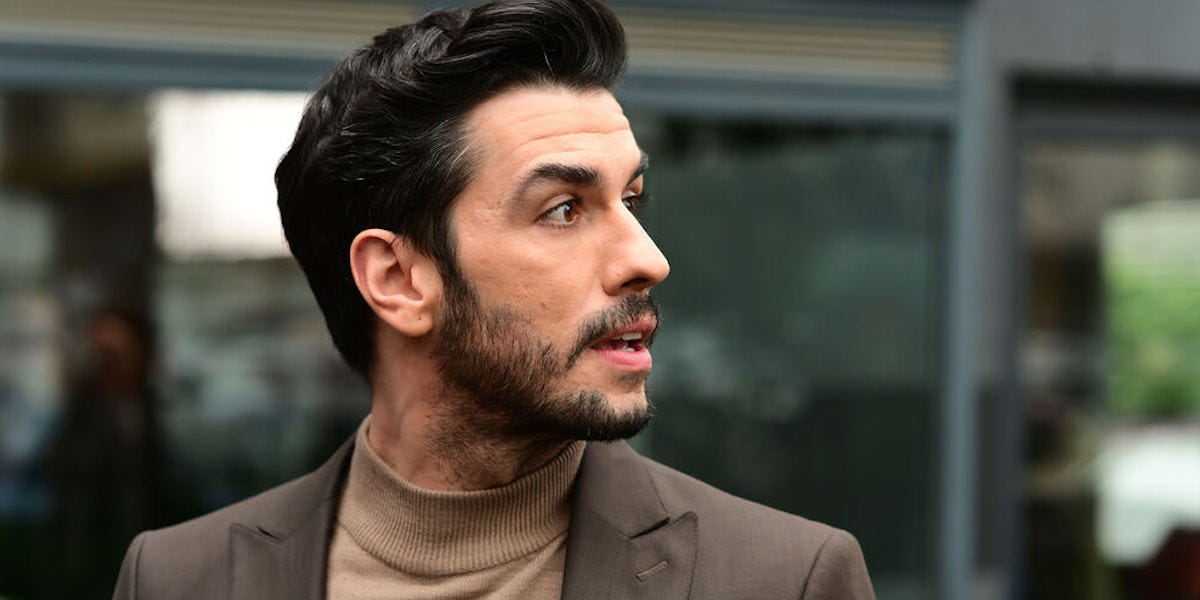 Aras Aydın'ın profil açısından çekilmiş portresi. Açık kahverengi balıkçı yaka kazak ve koyu renk bir ceket giymiş. Hafif sakalları var ve yüzünde düşünceli bir ifadeyle kadrajın dışına bakıyor. Arka plan bulanık, bir iç mekân görüntüsü.