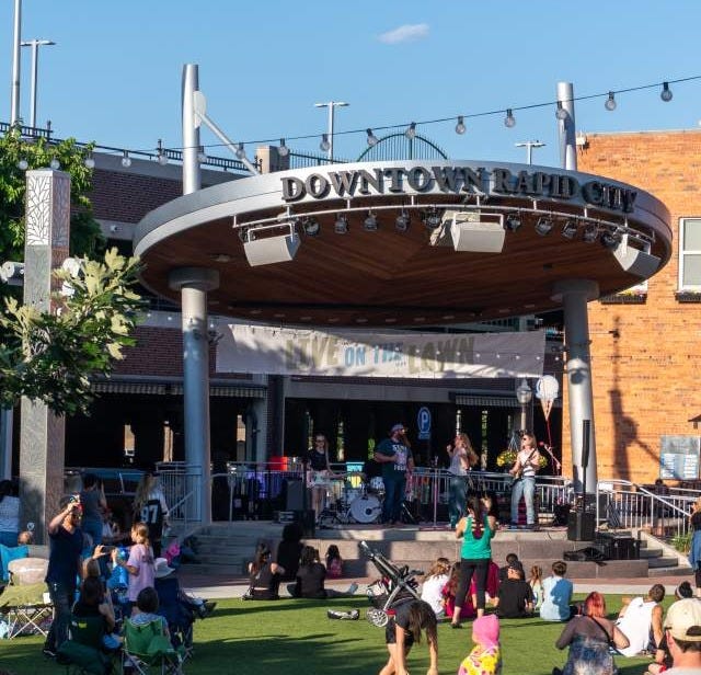 Looking at a distance towards a band playing on the stage at Main Street Square while attendees sit in lawn chairs scattered throughout the grass. Looking at a distance towards a band playing on the stage at Main Street Square while attendees sit in lawn chairs scattered throughout the grass.
