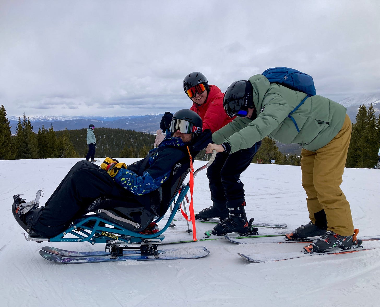Ben sits in a sit ski with two friends on skis behind him. Ben sits in a sit ski with two friends on skis behind him.