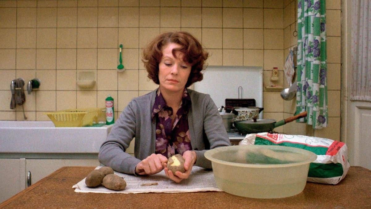 A still from Chantal Akerman’s 1975 movie Jeanne Dielman, 23, quai du Commerce, 1080 Bruxelles. Jeanne, a middle-aged woman with short fluffy brown hair, is sitting dejectedly in a small kitchen peeling potatoes.