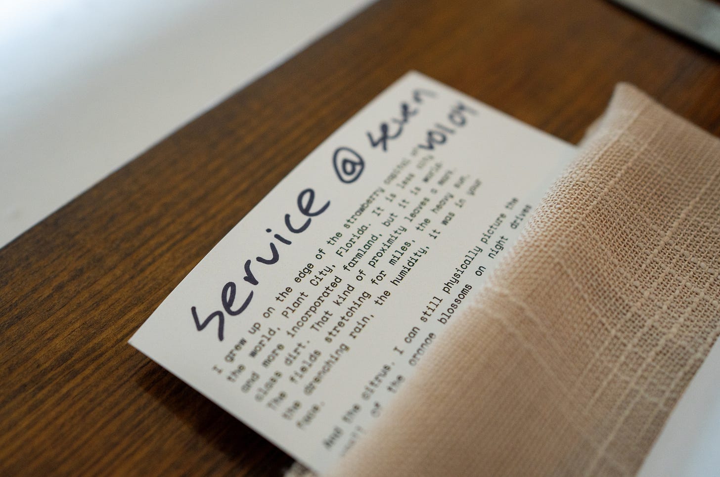 A handwritten welcome note for Service at Seven Volume 4 resting on a wooden table next to a folded tan linen napkin.