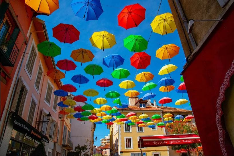 Umbrellas of many bright colors hang suspended over a courtyard surrounded by brightly colored buildings on a clear, sunny day Umbrellas of many bright colors hang suspended over a courtyard surrounded by brightly colored buildings on a clear, sunny day