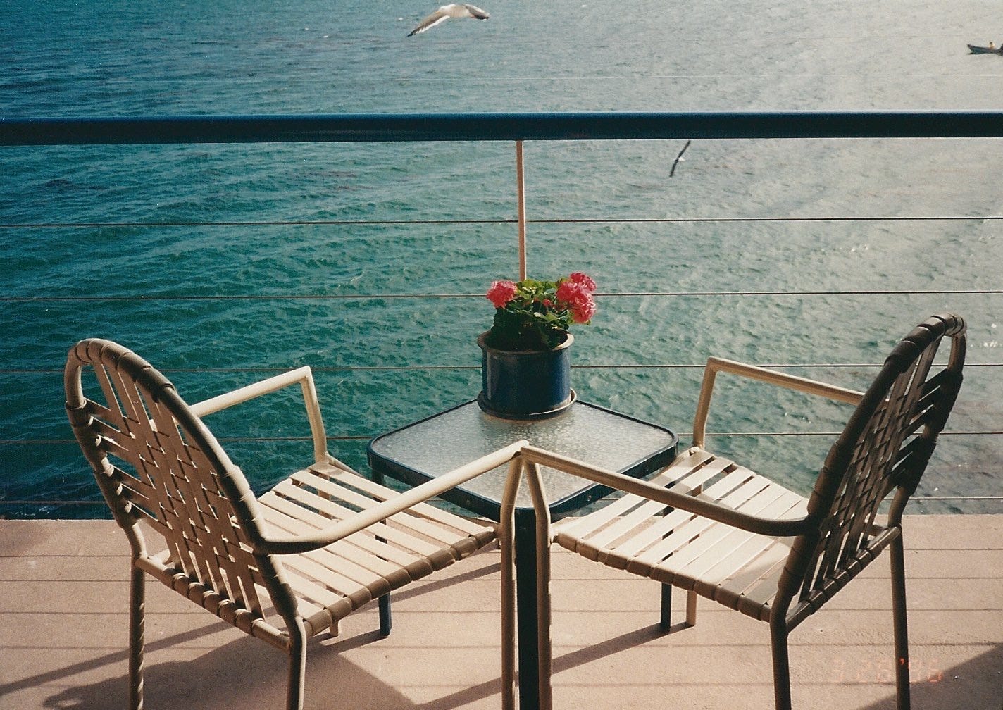 Two empty patio chairs and a small table with flowers on a waterfront deck, photographed on film. Two empty patio chairs and a small table with flowers on a waterfront deck, photographed on film.