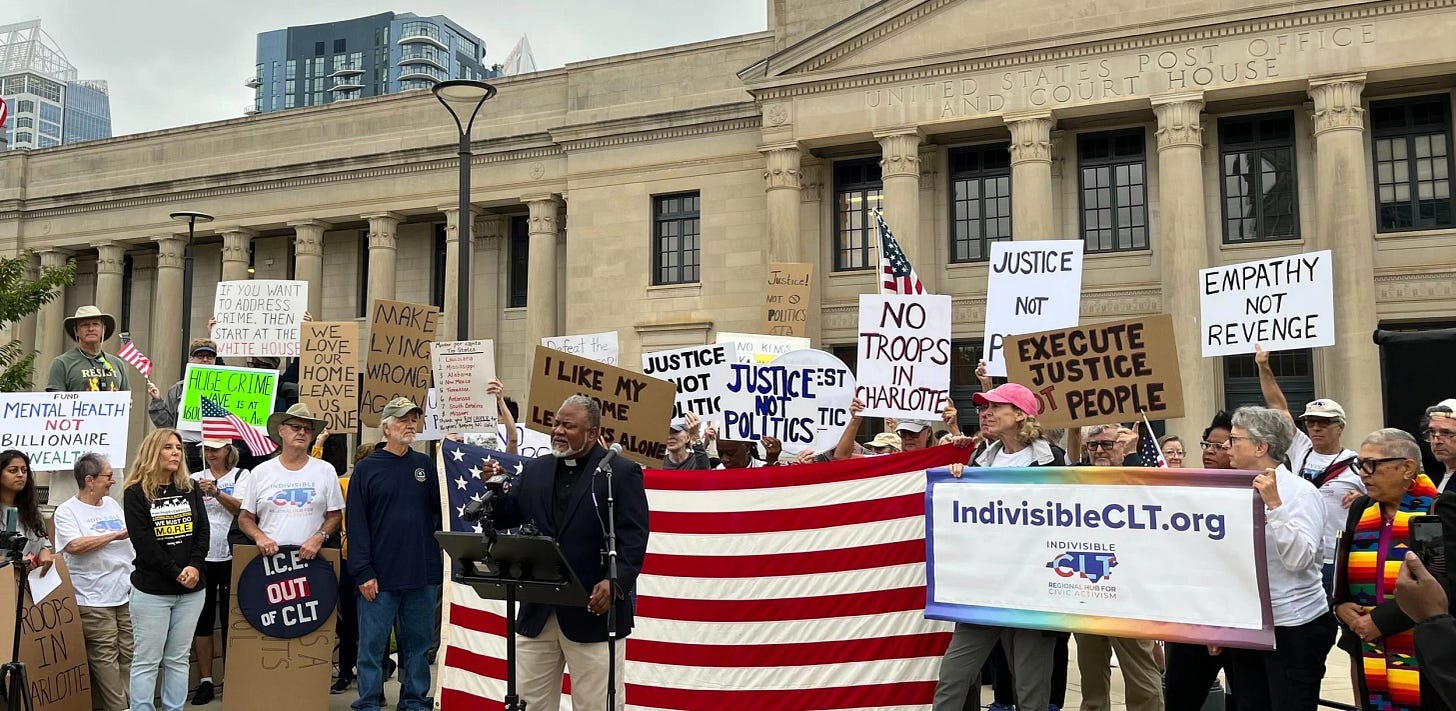 A diverse group of protesters gathers in front of a neoclassical U.S. Post Office and Court House building in Charlotte, holding handmade signs with messages like “Justice Not Politics,” “No Troops in Charlotte,” and “Empathy Not Revenge,” while a speaker stands at a podium in front of a large U.S. flag and an “IndivisibleCLT.org” banner.