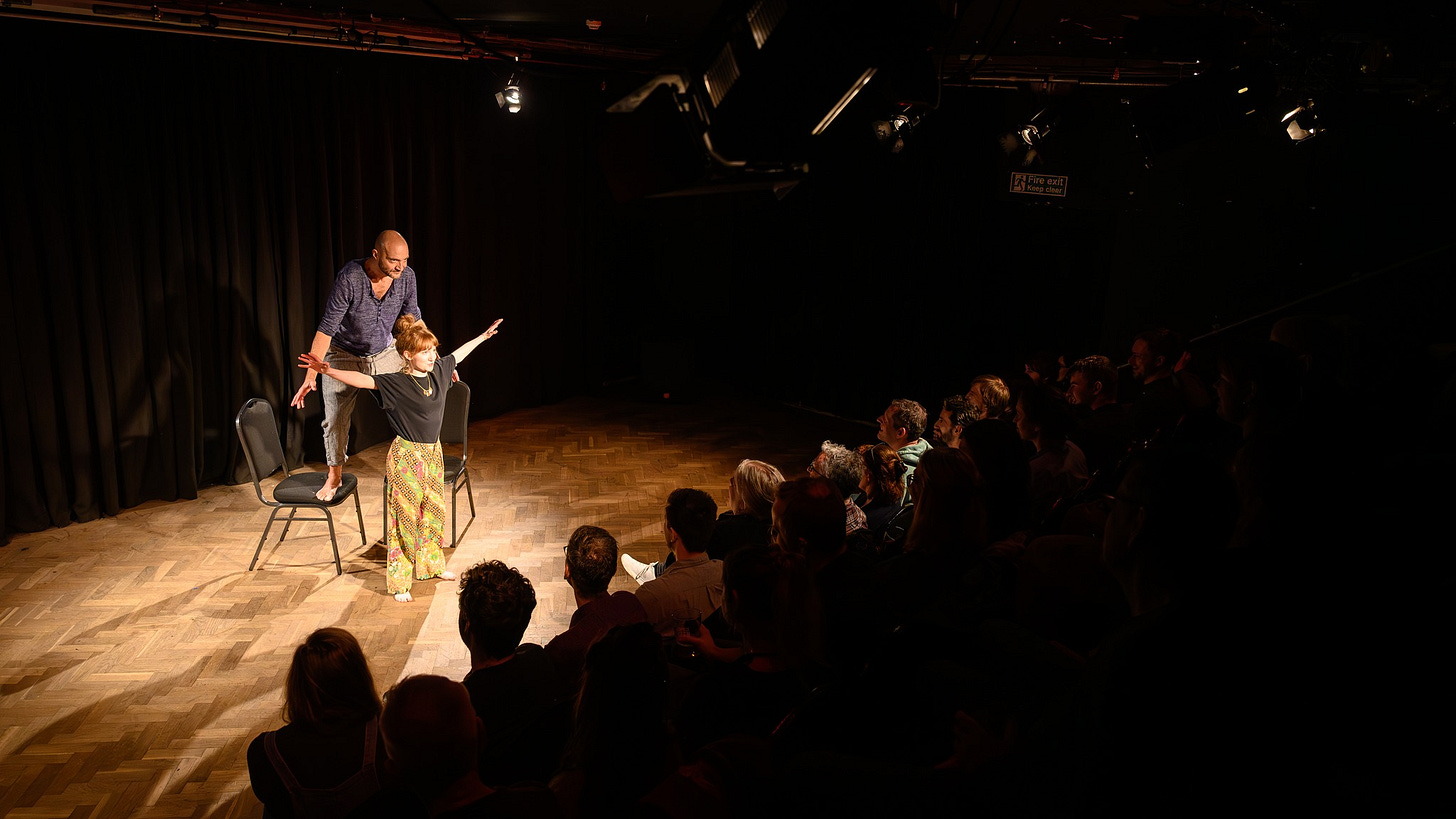 Two performers stand on stage. A woman with red hair stands with her arms out and a man in a blue top stands on two chairs behind her