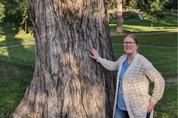 A white woman stands next to a giant tree in a park