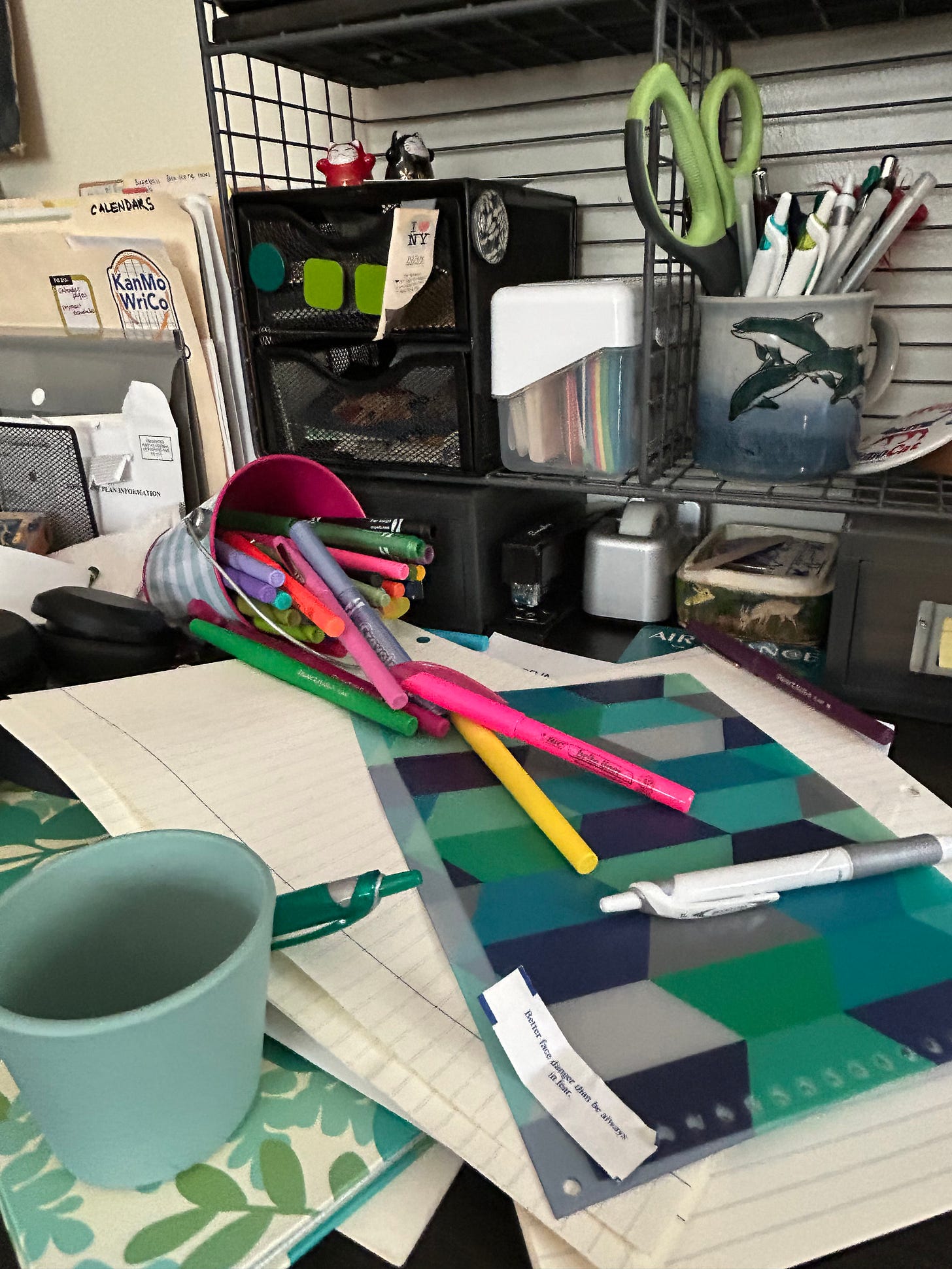 piles of paper, notebooks and other stuff on a desk: a small cup turned over, pens and colorful magic markers, a steel shelf crowded with stuff.