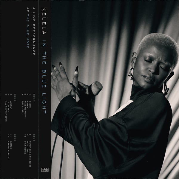 Album cover for In The Blue Light. It is a black and white photo of Kelela, a young black woman with a shaved head and long, painted fingernails, who is wearing a black dress with long sleeves and is holding a microphone between her hands as she closes her eyes. She is standing in front of a curtain backdrop.