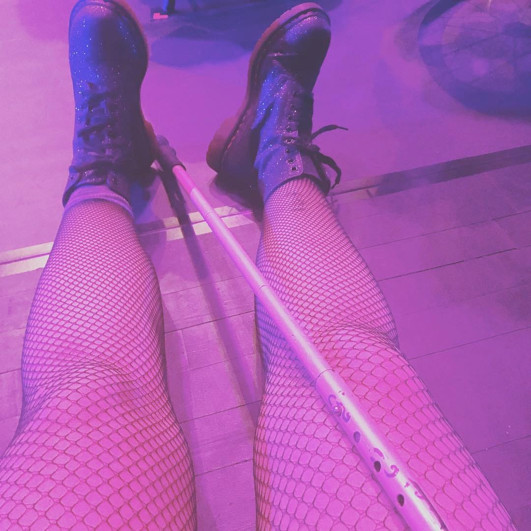 A square frame captures a medium close-up of someone sitting on the floor, Two legs adorned in delicate fishnet stockings rest gently, while sparkling rubber boots ignite a sense of daring, joy, and rebellion. Between them, a metal cane rest on their legs.
